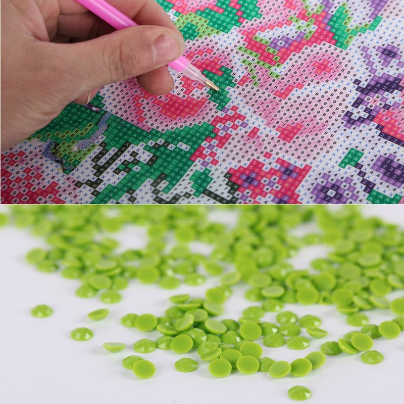 Diamond painting kit with a hand using a pink tool to place green rhinestones onto a canvas with printed symbols, and a pile of loose green rhinestones.