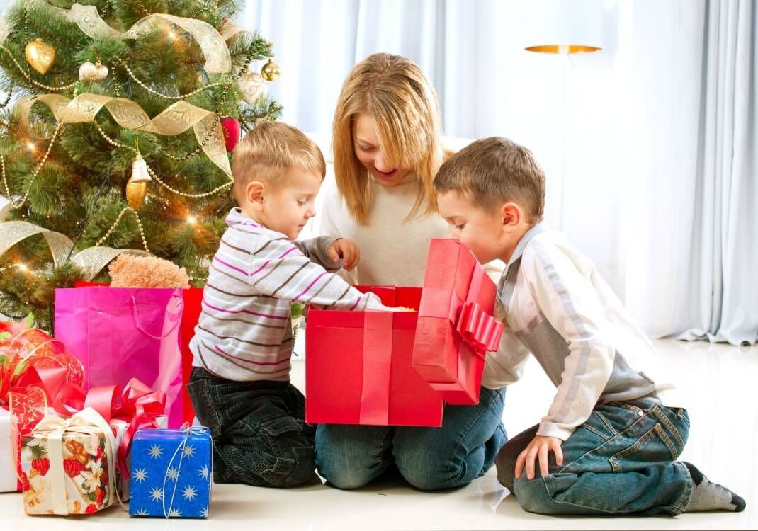 Mother and two sons opening a large red gift box next to a decorated Christmas tree with presents.