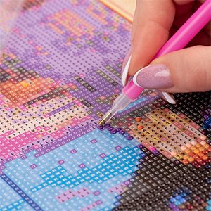 Close-up of a person's hand using a pink tool to place a diamond onto a colorful 5D diamond painting kit with a beach lighthouse design.