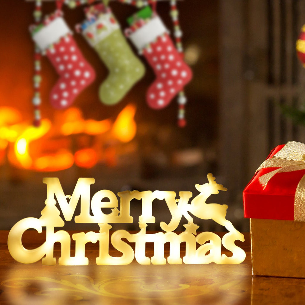 Lighted 'Merry Christmas' sign with reindeer and tree accents, next to a gift box, with stockings and fireplace in background.