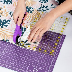 Hands using a purple rotary cutter and acrylic ruler to cut patterned fabric on a purple self-healing mat.
