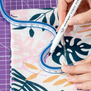 Hands using a flexible ruler and marker to draw on floral fabric, with a purple cutting mat visible.