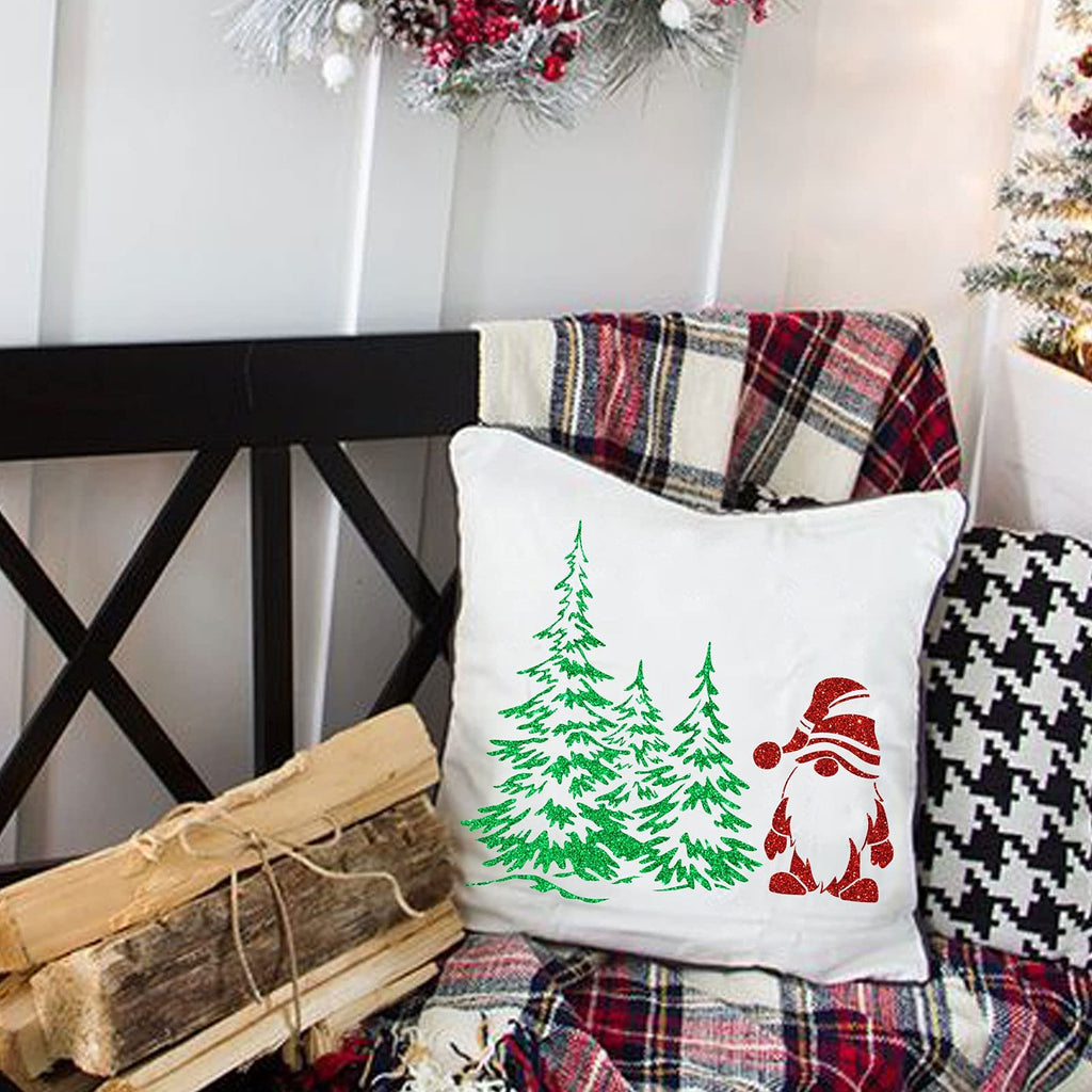 White pillow with green glitter Christmas trees and a red glitter gnome design, set against a festive backdrop with plaid blankets and logs.