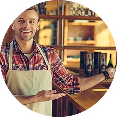 Smiling man in apron and plaid shirt gesturing towards a bar counter with shelves of bottles behind him