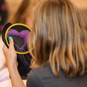 Person's hands holding a black embroidery hoop with a purple heart cross-stitch pattern.