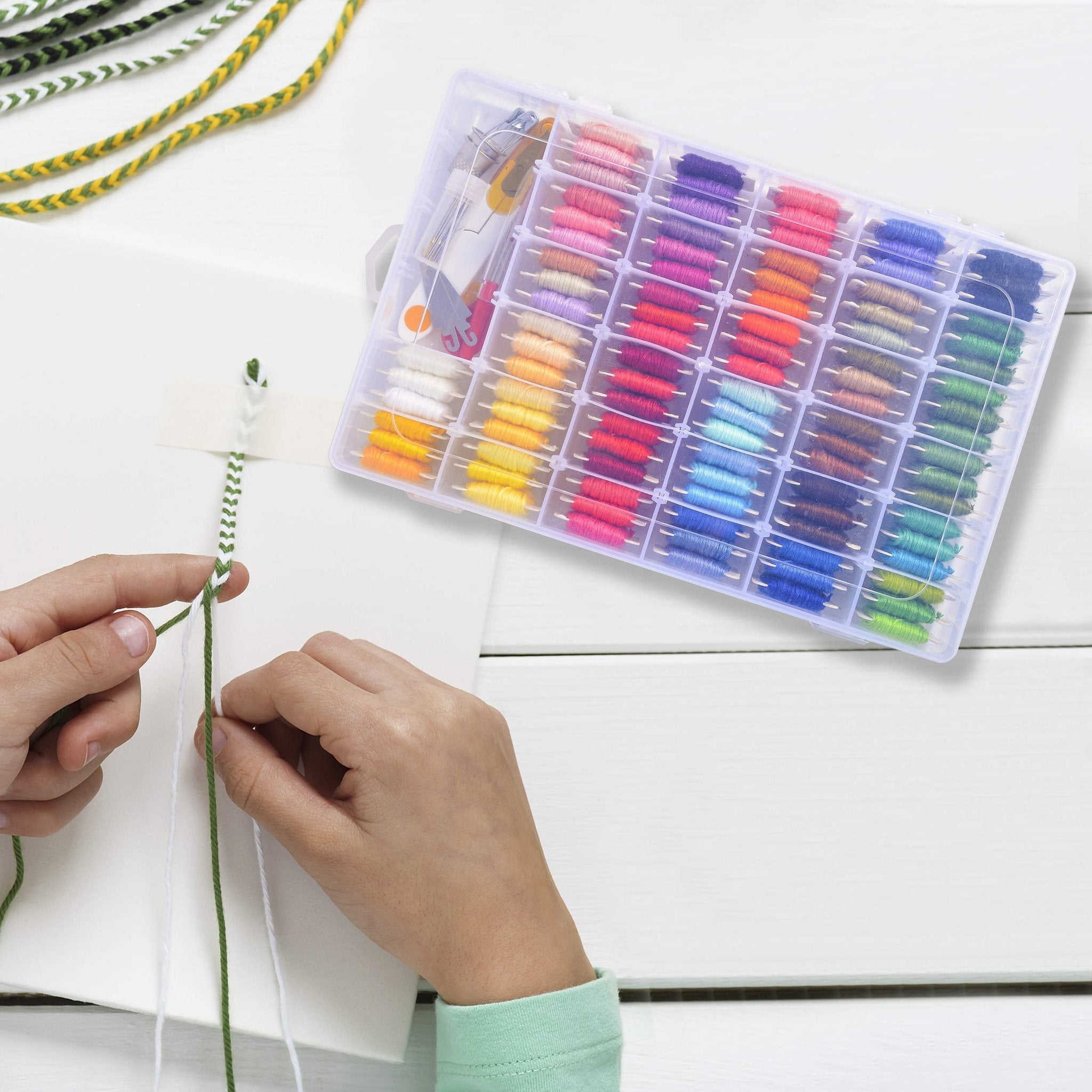 Hands braiding green and white thread next to a clear organizer box filled with colorful embroidery floss.
