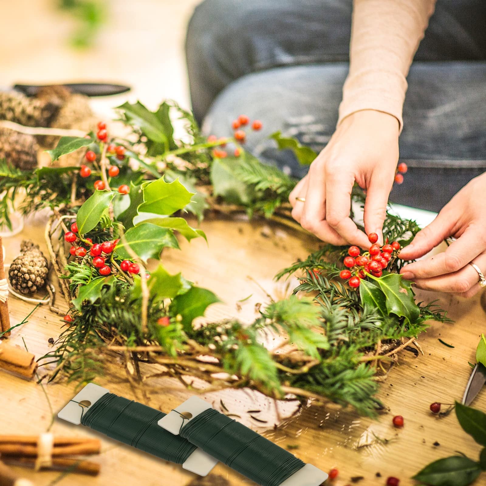 Hands crafting a wreath with greenery and red berries, two spools of green floral wire in foreground.