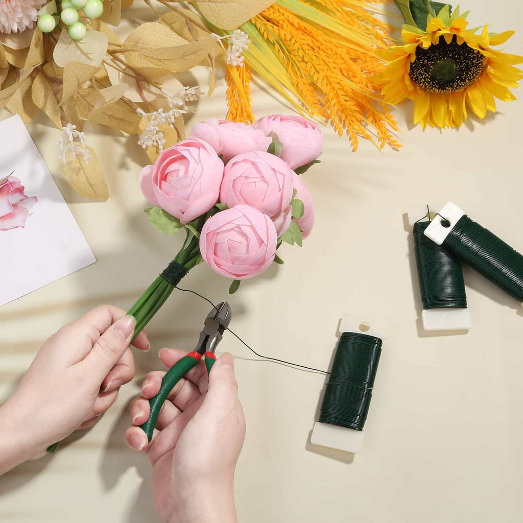 Hands using wire cutters to trim green floral wire from a spool, with pink artificial flowers and decorative foliage in the background.