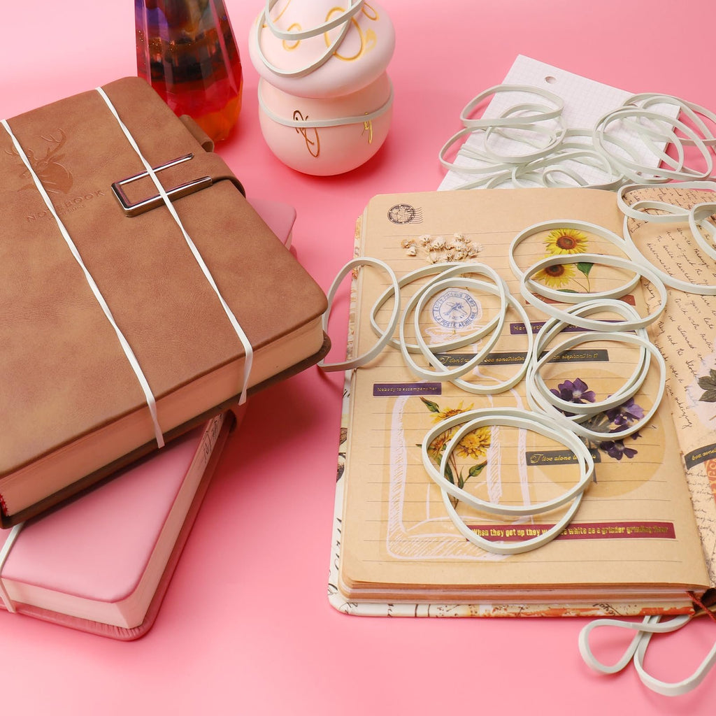 Light green rubber bands scattered on open notebooks with floral and vintage designs, alongside decorative pink and brown notebooks.
