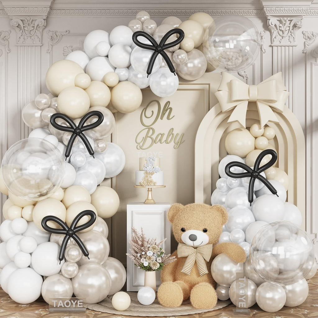 Pearl white and cream balloon arch with clear balloons, black bows, a teddy bear, and a cake for a baby shower.