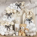 Pearl white and cream balloon arch with clear balloons, black bows, a teddy bear, and a cake for a baby shower.