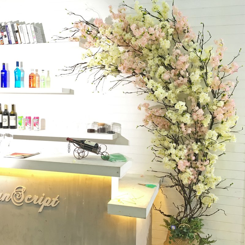 Artificial cherry blossom tree branches with pink and cream flowers against a white wall with shelves of bottles and books.