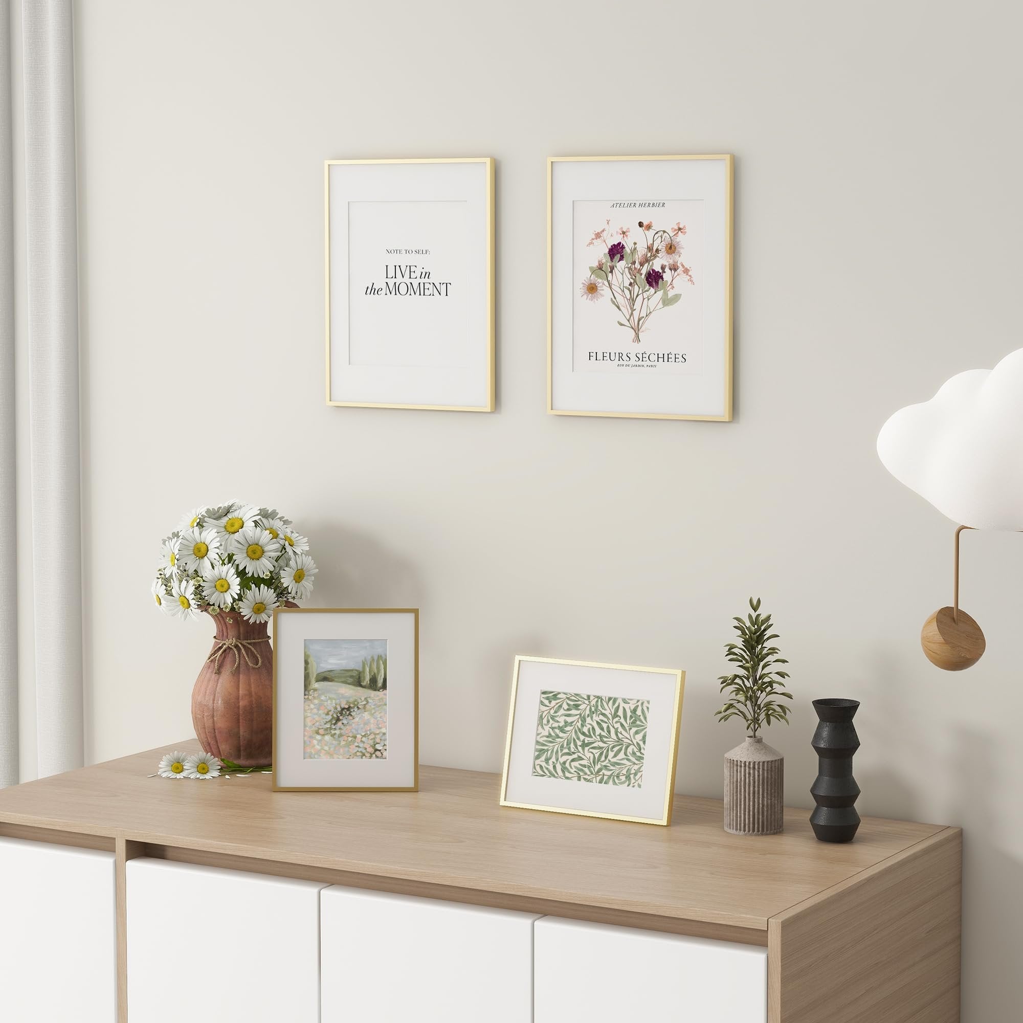 Two gold picture frames on a wall above a wooden cabinet with decorative items, one frame contains text and the other a floral illustration.