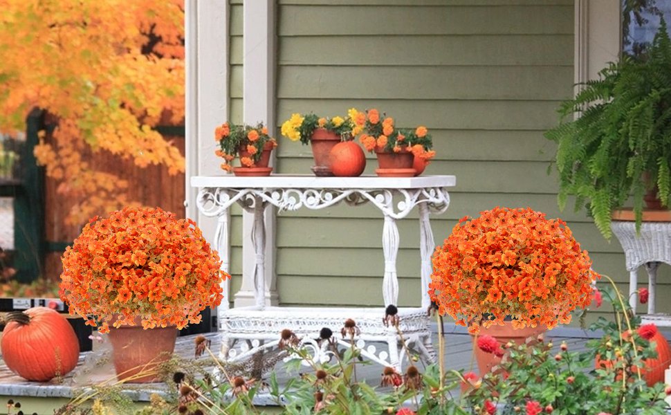 Two potted orange mum bushes flank a white table on a porch with pumpkins and fall foliage