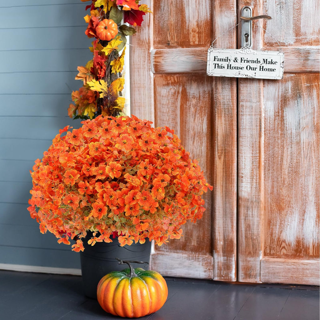 Orange artificial mums in a black pot with a pumpkin and fall garland on a porch.