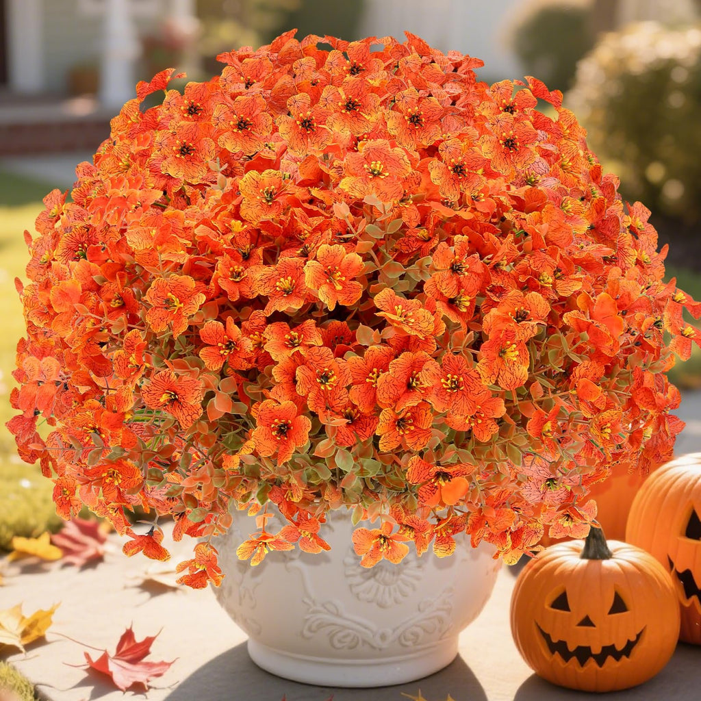 Artificial orange mums with eucalyptus leaves in a white pot, with pumpkins and fall leaves on a patio.