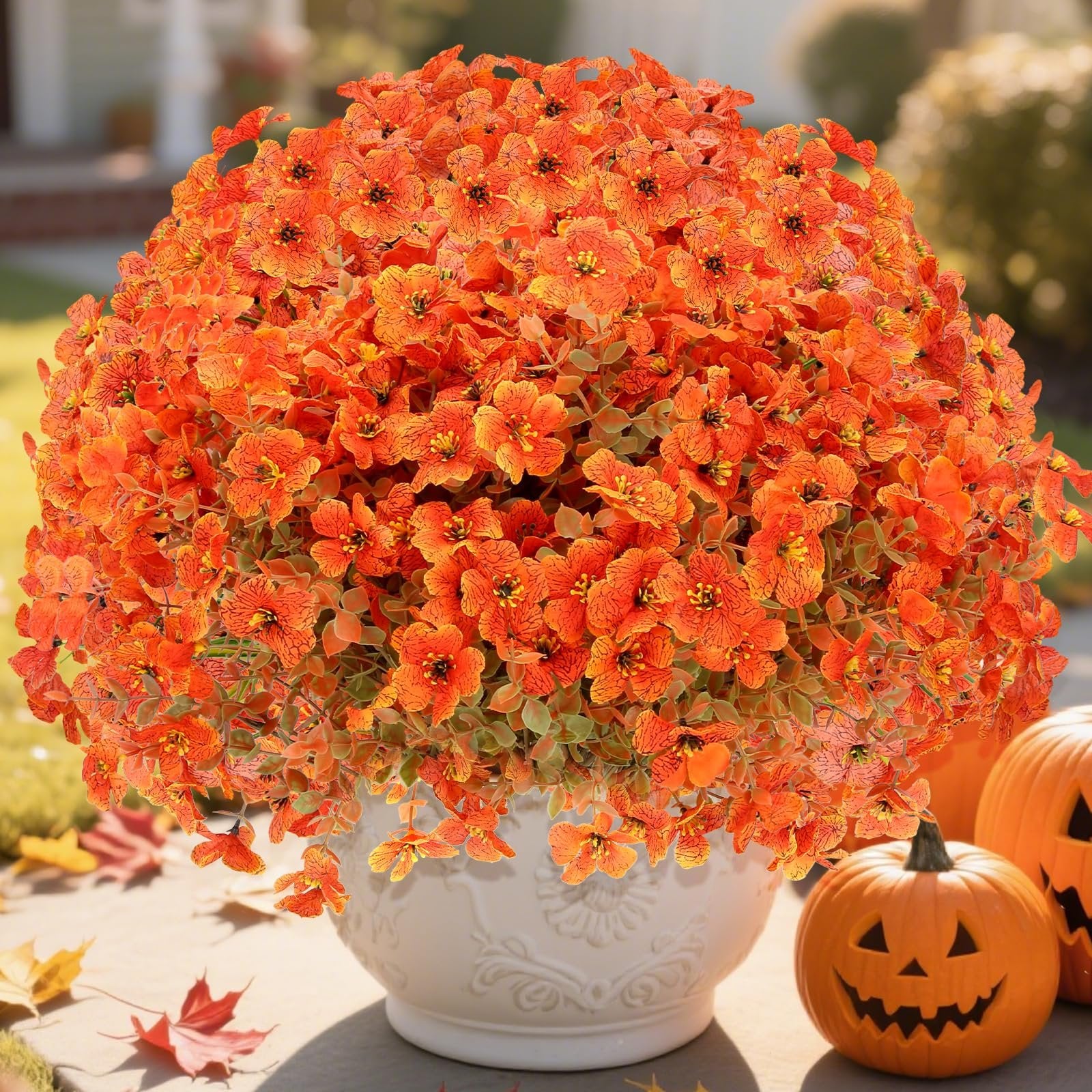Artificial orange mums with eucalyptus leaves in a white pot, with pumpkins and fall leaves on a patio.
