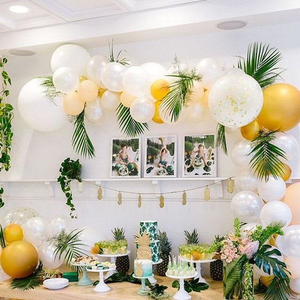 Tropical party setup with balloon garland, fruit platters, cake, and framed photos on a shelf.