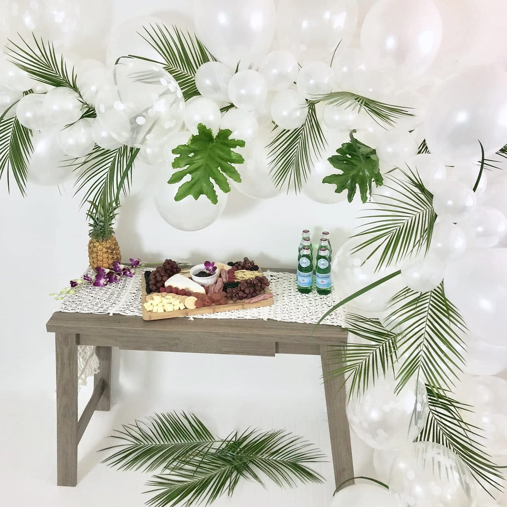 Tropical party table setup with charcuterie board, pineapple, and white balloons decorated with palm leaves.