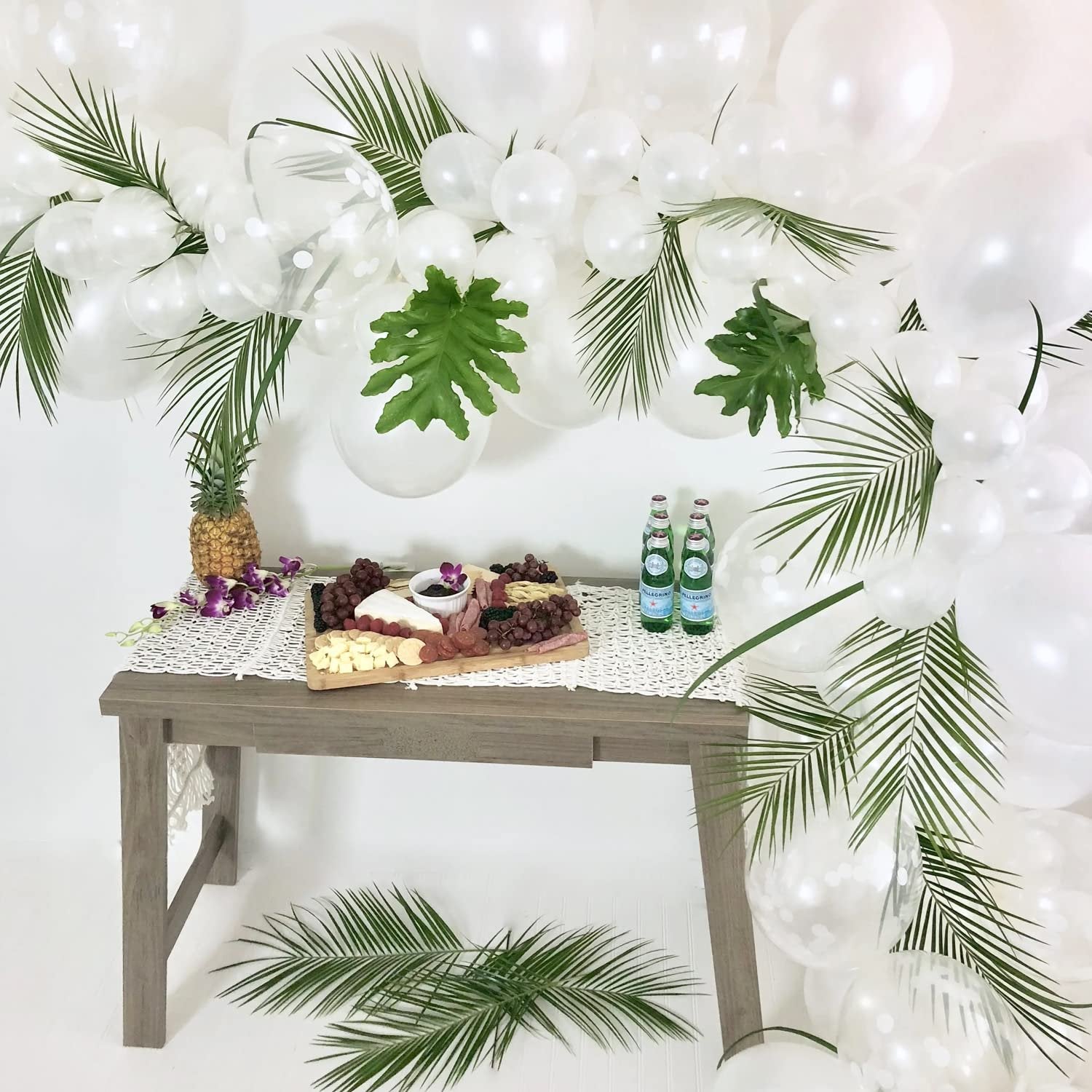 Tropical party table setup with charcuterie board, pineapple, and white balloons decorated with palm leaves.