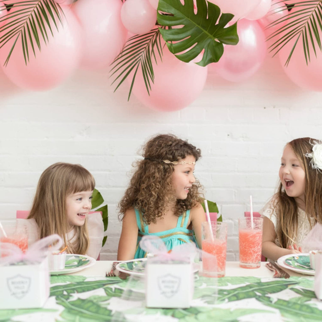 Three young girls at a party table decorated with pink balloons and tropical leaves.