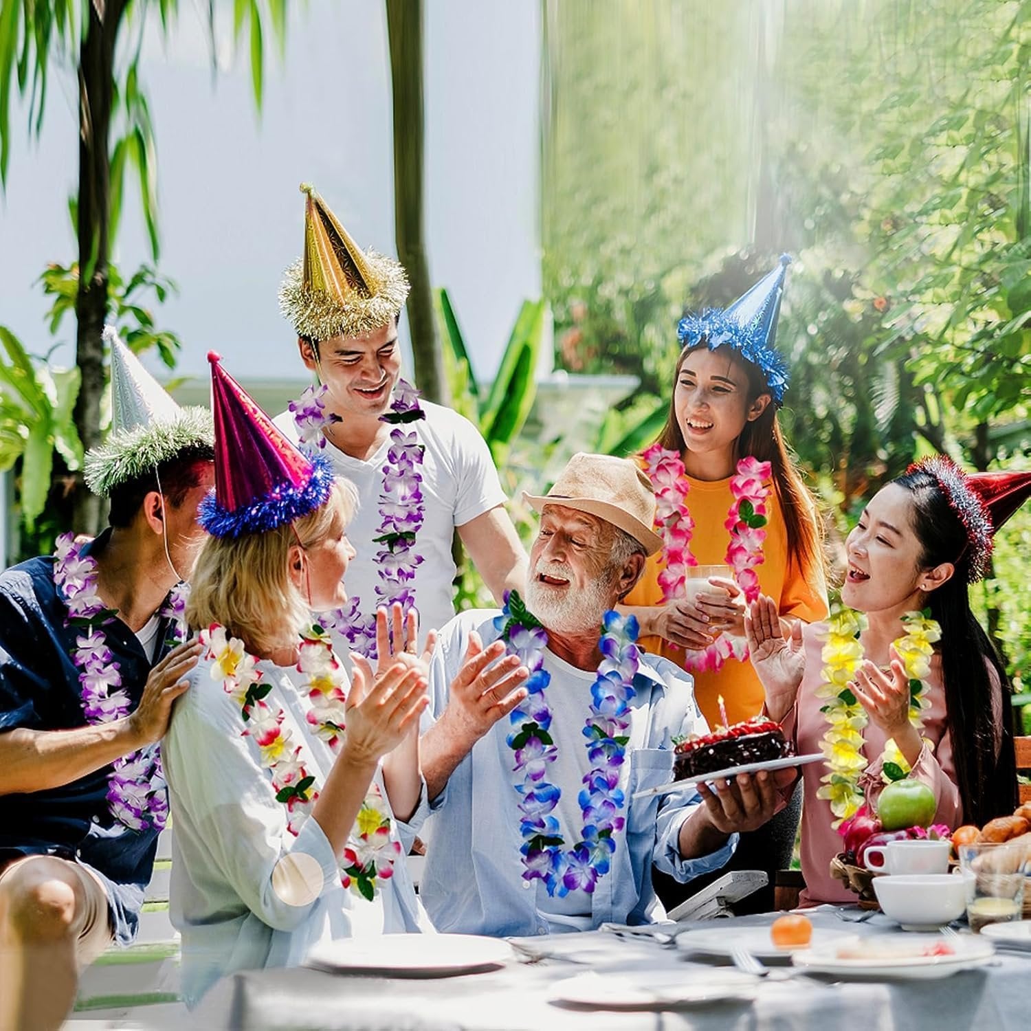 Group of people celebrating outdoors wearing party hats and flower leis, with a birthday cake on the table.