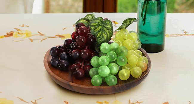 Three bunches of artificial grapes in red, green, and yellow on a wooden plate, with a green vase in the background.
