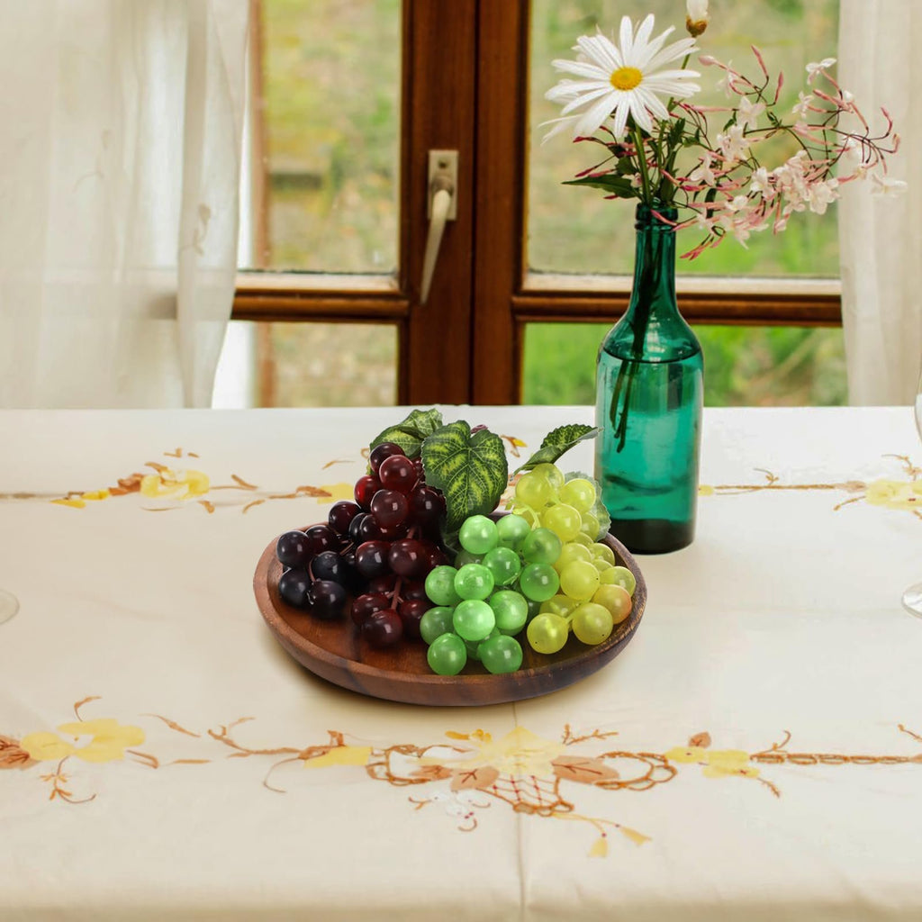 Three bunches of artificial grapes in red, dark purple, and green on a wooden plate, with a vase of flowers in the background.