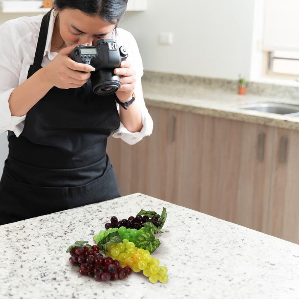 Woman in apron photographing artificial grapes in various colors on a speckled countertop