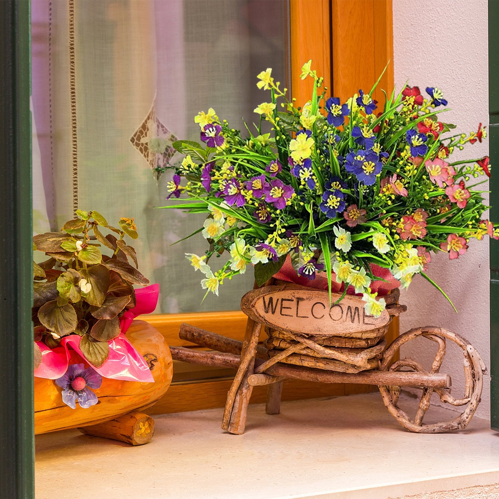 Artificial flowers in a rustic wooden wheelbarrow planter with a welcome sign, next to a potted plant.