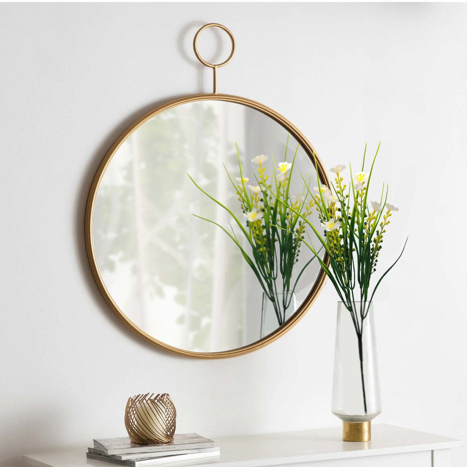 Round gold-framed mirror hanging on a white wall above a white shelf with decorative items.
