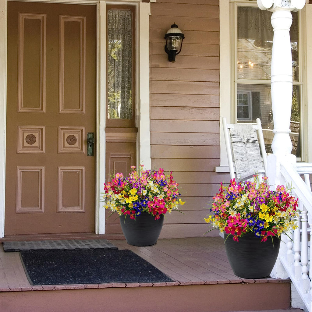 Two black planters filled with colorful artificial flowers on a wooden porch next to a brown door.