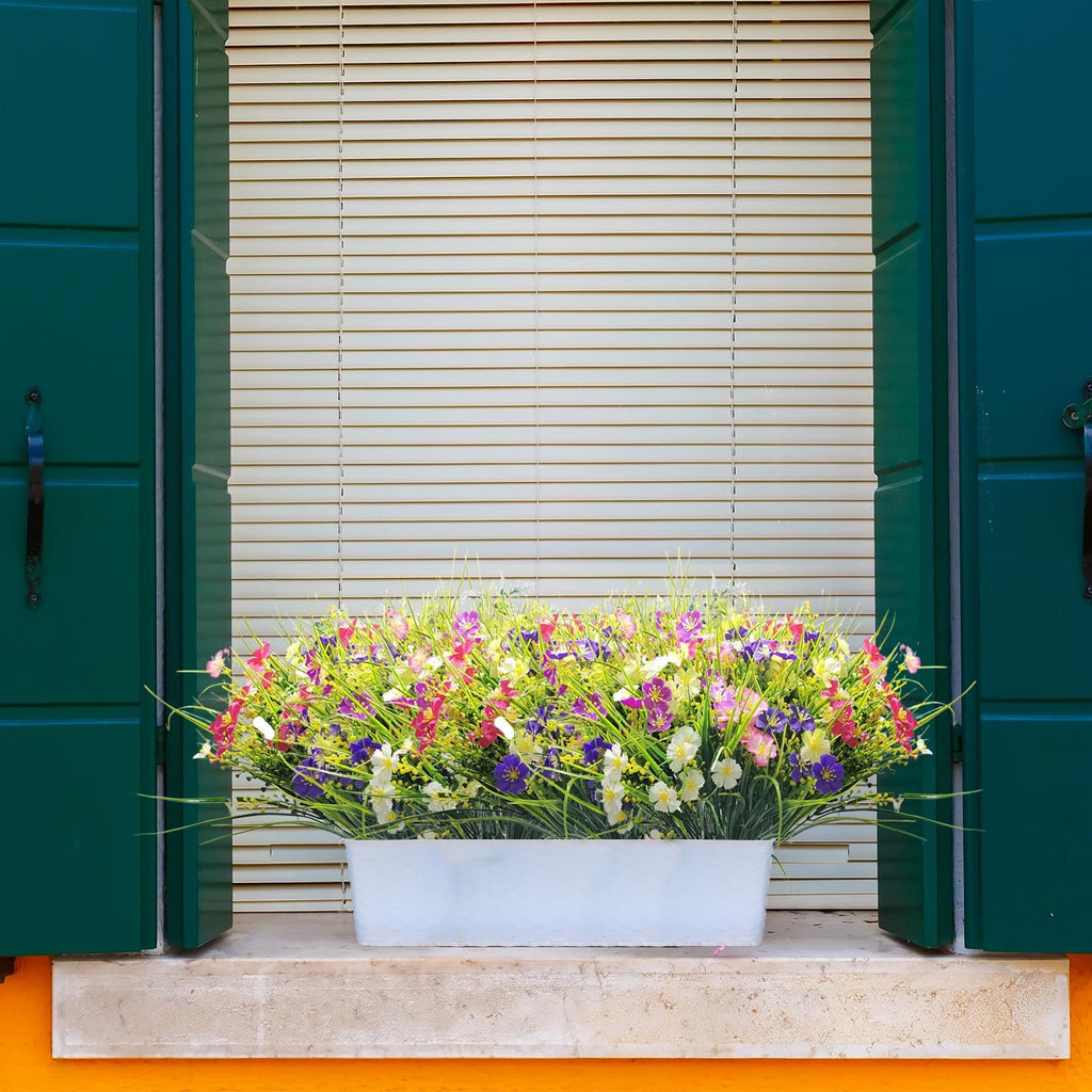 Artificial flowers in a white planter box on a windowsill with green shutters and blinds