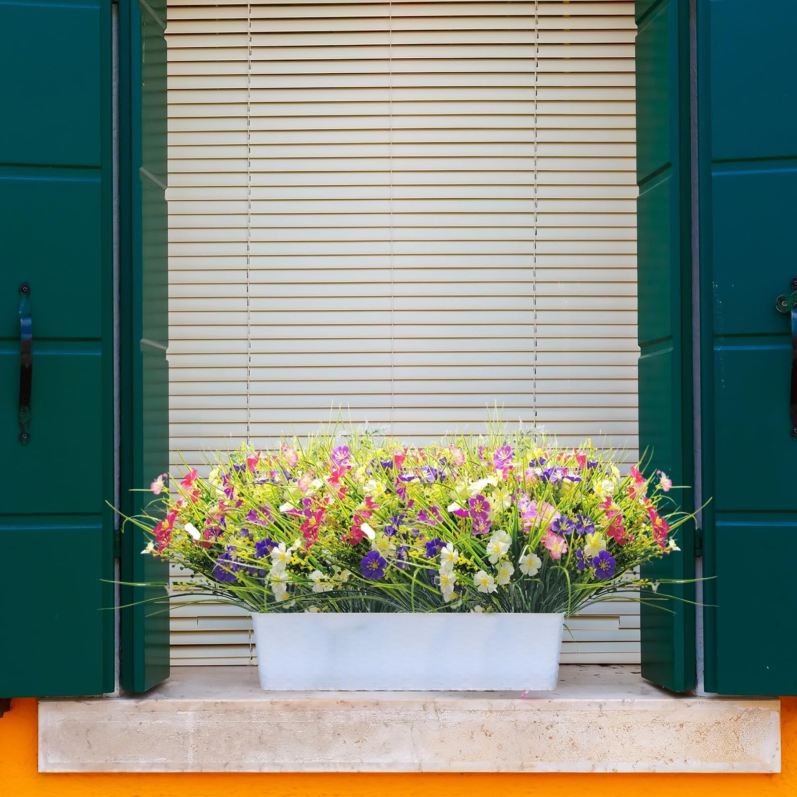 Artificial flowers in a white planter box on a windowsill with green shutters and blinds