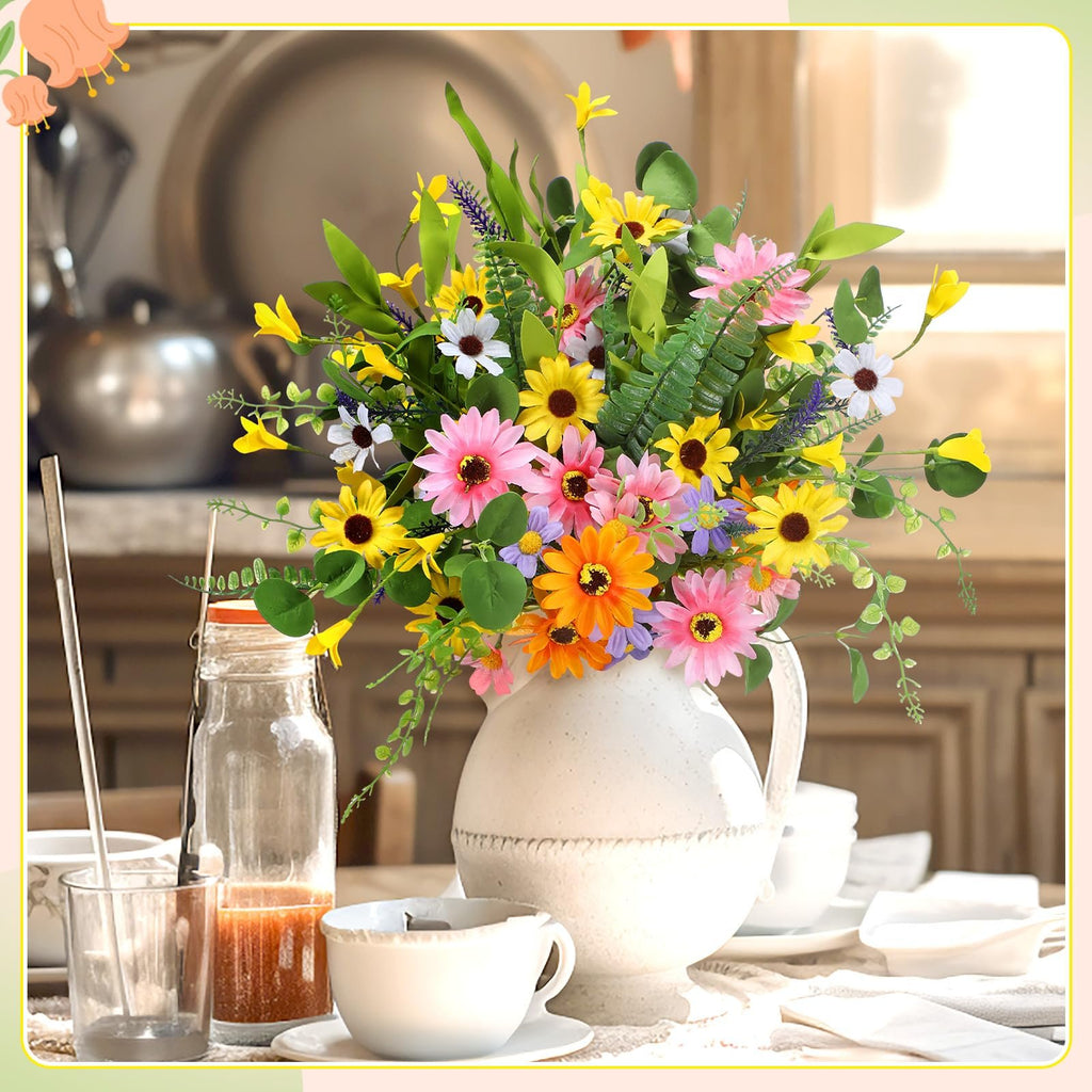 Colorful artificial daisy wildflowers in a white pitcher on a table with dishes and a glass bottle.