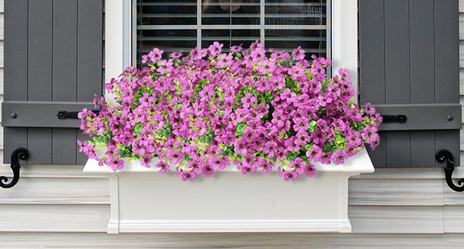 Window box filled with vibrant purple artificial flowers and green foliage, framed by dark gray shutters.