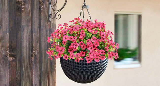 Hanging basket filled with vibrant pink artificial daisy flowers and green foliage, suspended by a chain against a wooden post and building.