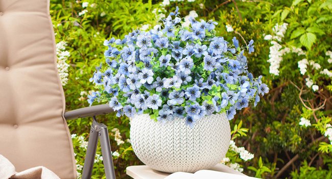 Bouquet of blue artificial flowers in a white textured pot on a table next to a chair, with lush green foliage in the background.