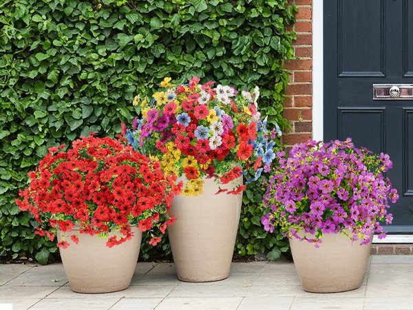 Three pots of artificial flowers in red, multicolor, and purple, arranged on a patio in front of a brick wall and dark door.
