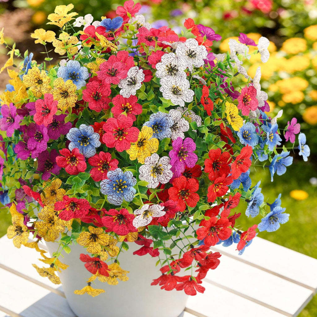 Bundle of multicolor artificial daisy flowers in a white pot on a white table, with a blurred garden background.
