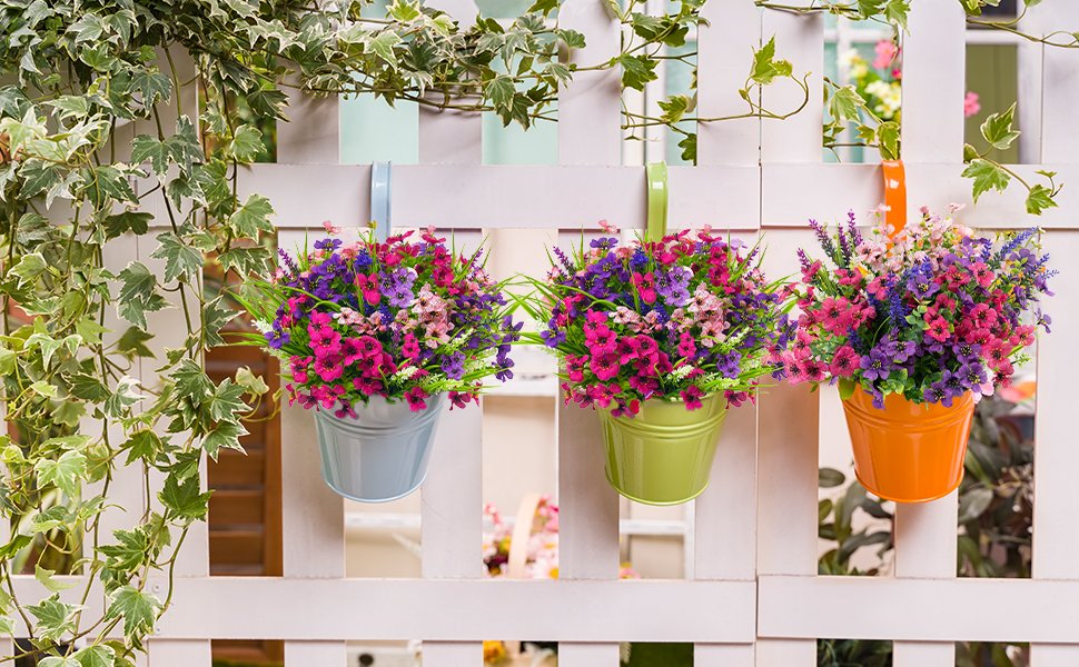 Three hanging buckets filled with colorful artificial flowers against a white fence and ivy.