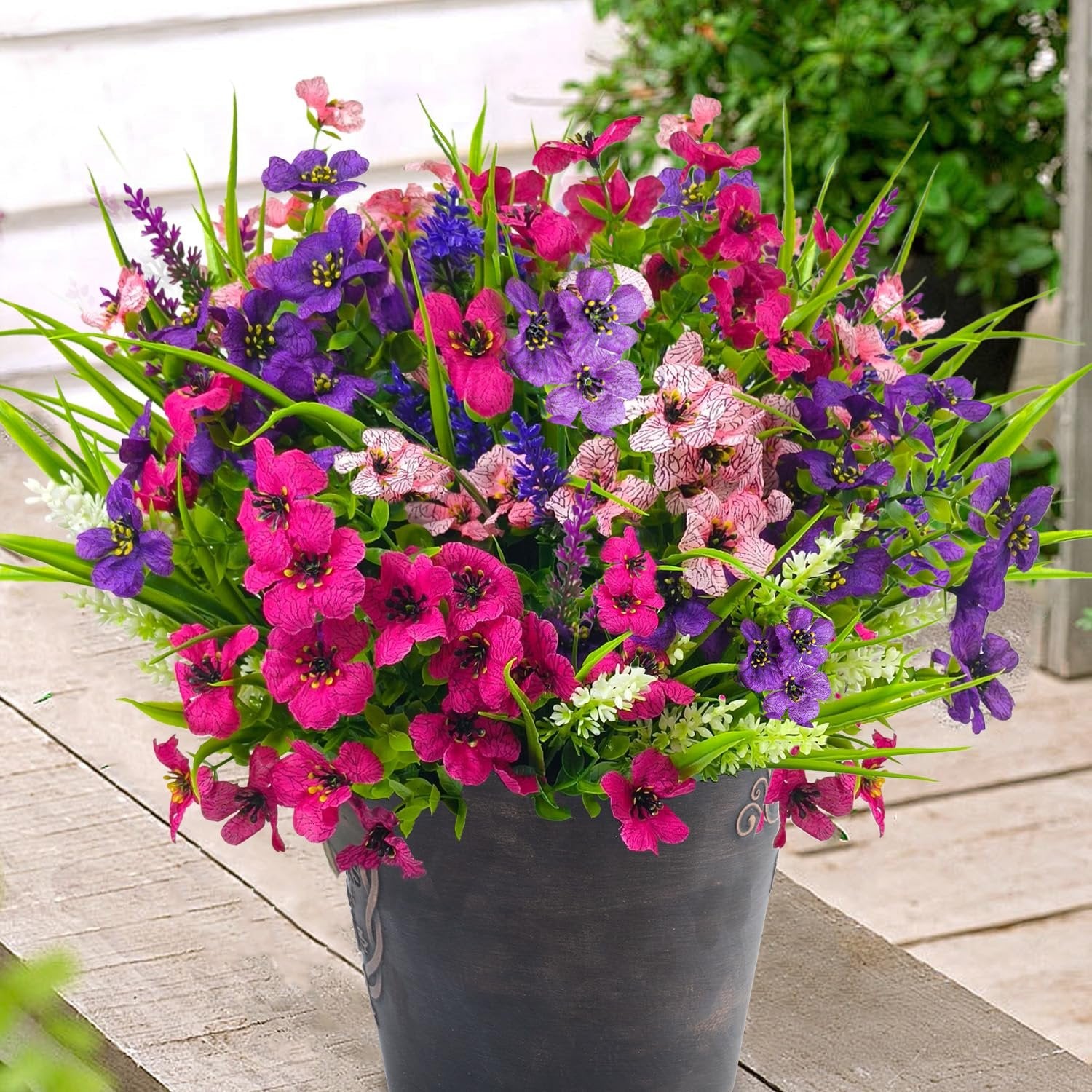 Mixed artificial flowers in a dark pot on a wooden surface, featuring pink, purple, and blue blooms with green foliage.