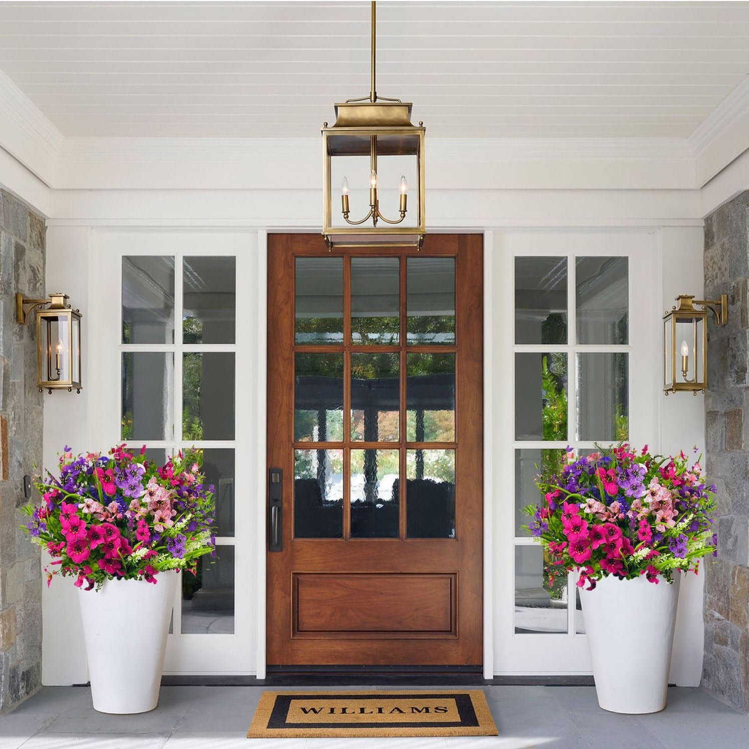 Two large white planters filled with colorful artificial flowers flank a wooden front door with glass panes.