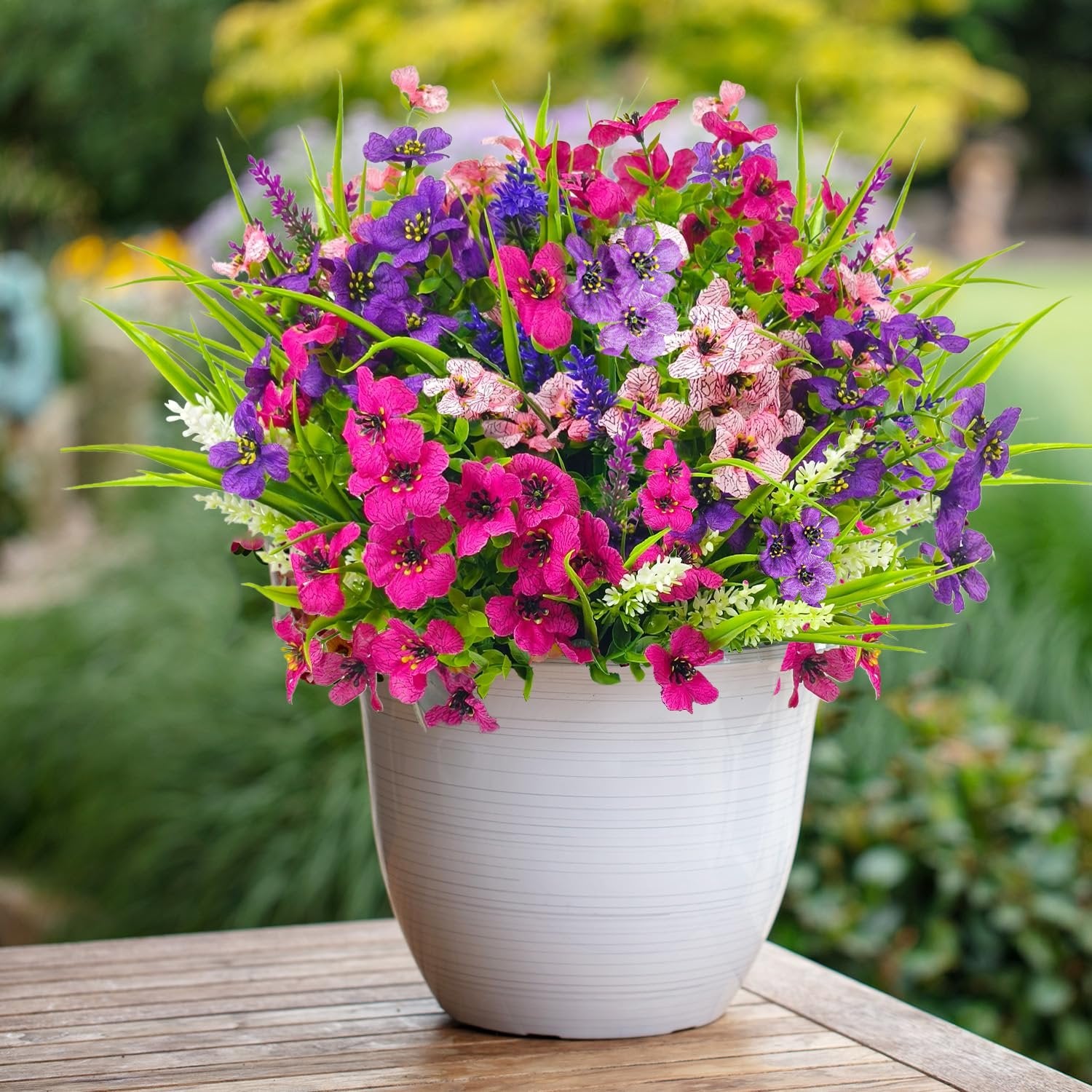 Mixed color artificial flowers in a white pot on a wooden table with a blurred garden background.
