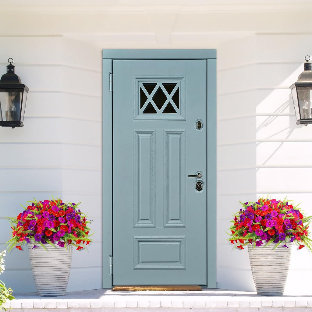 Two white planters filled with vibrant artificial petunia and fern flowers flank a light blue front door on a white porch.