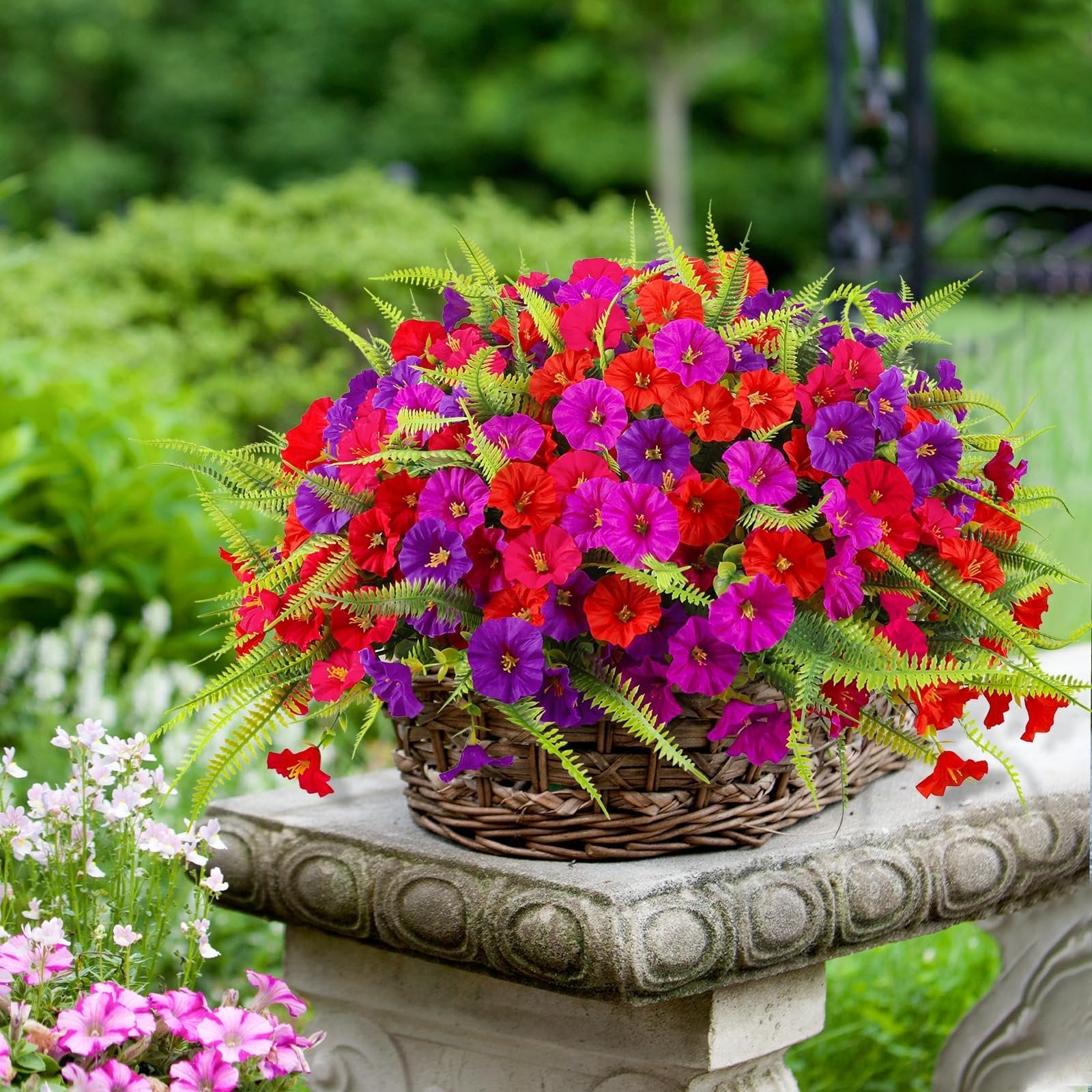 Wicker basket filled with artificial red, purple, and pink petunia and fern flowers, displayed outdoors on a stone pedestal.