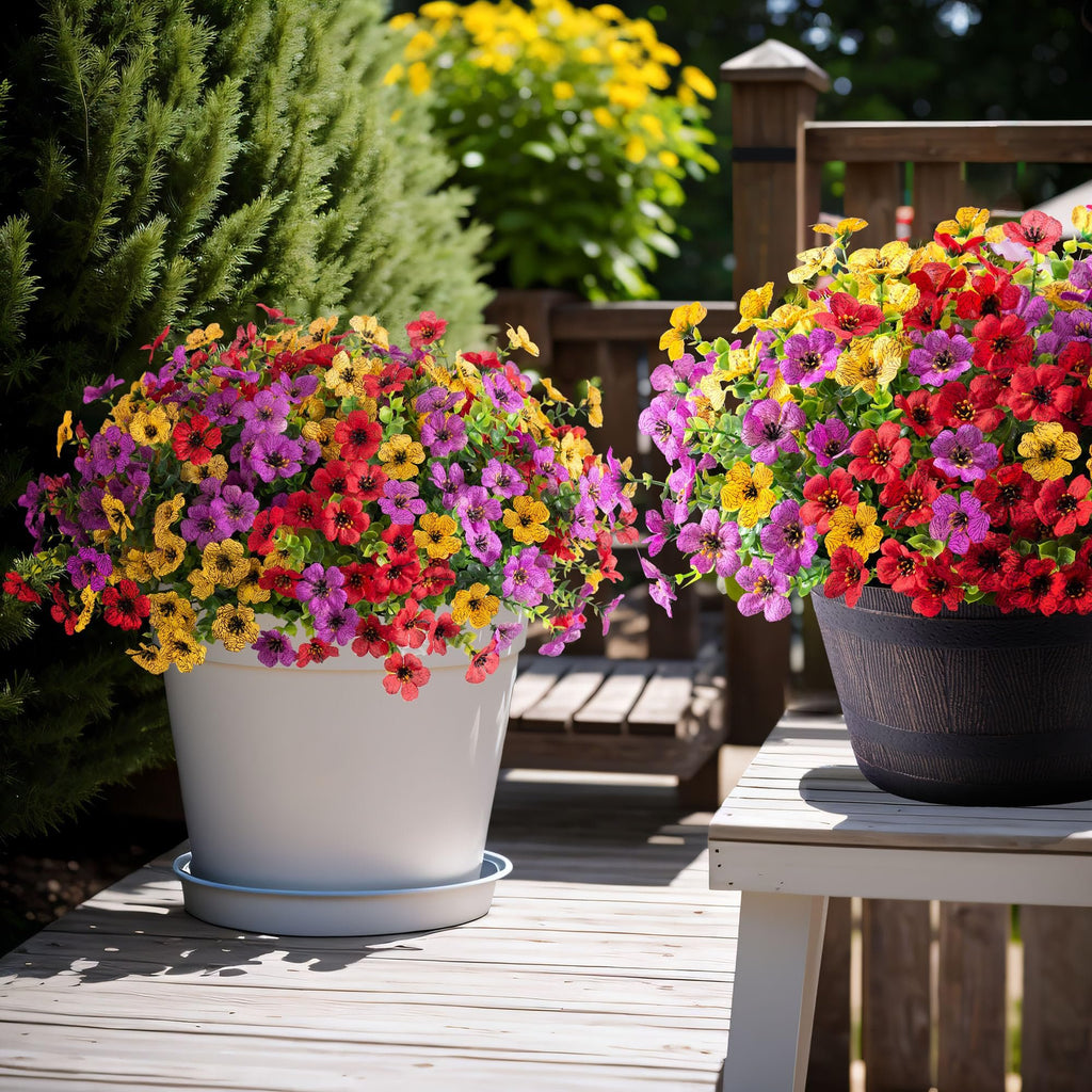 Two pots of colorful artificial daisy flowers with eucalyptus leaves on a wooden deck, one white pot and one dark pot.