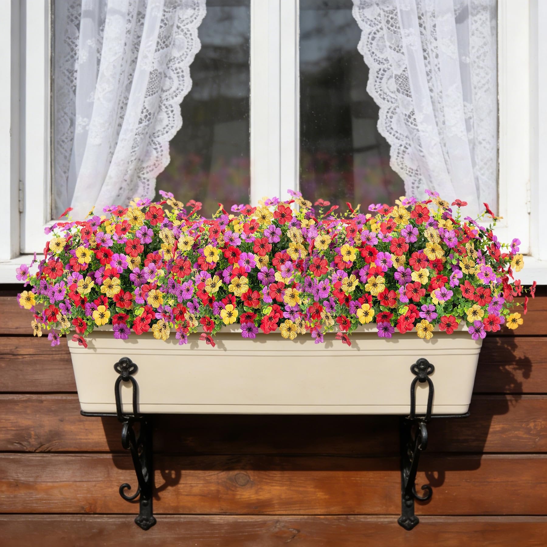 Window box filled with vibrant artificial daisy flowers and eucalyptus leaves, mounted on a wooden wall with lace curtains visible inside the window.