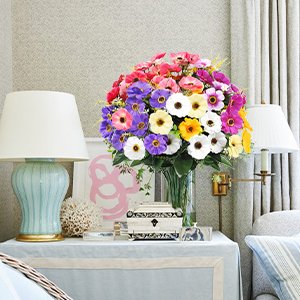 Vase of mixed color artificial daisy silk flowers on a table between two lamps.
