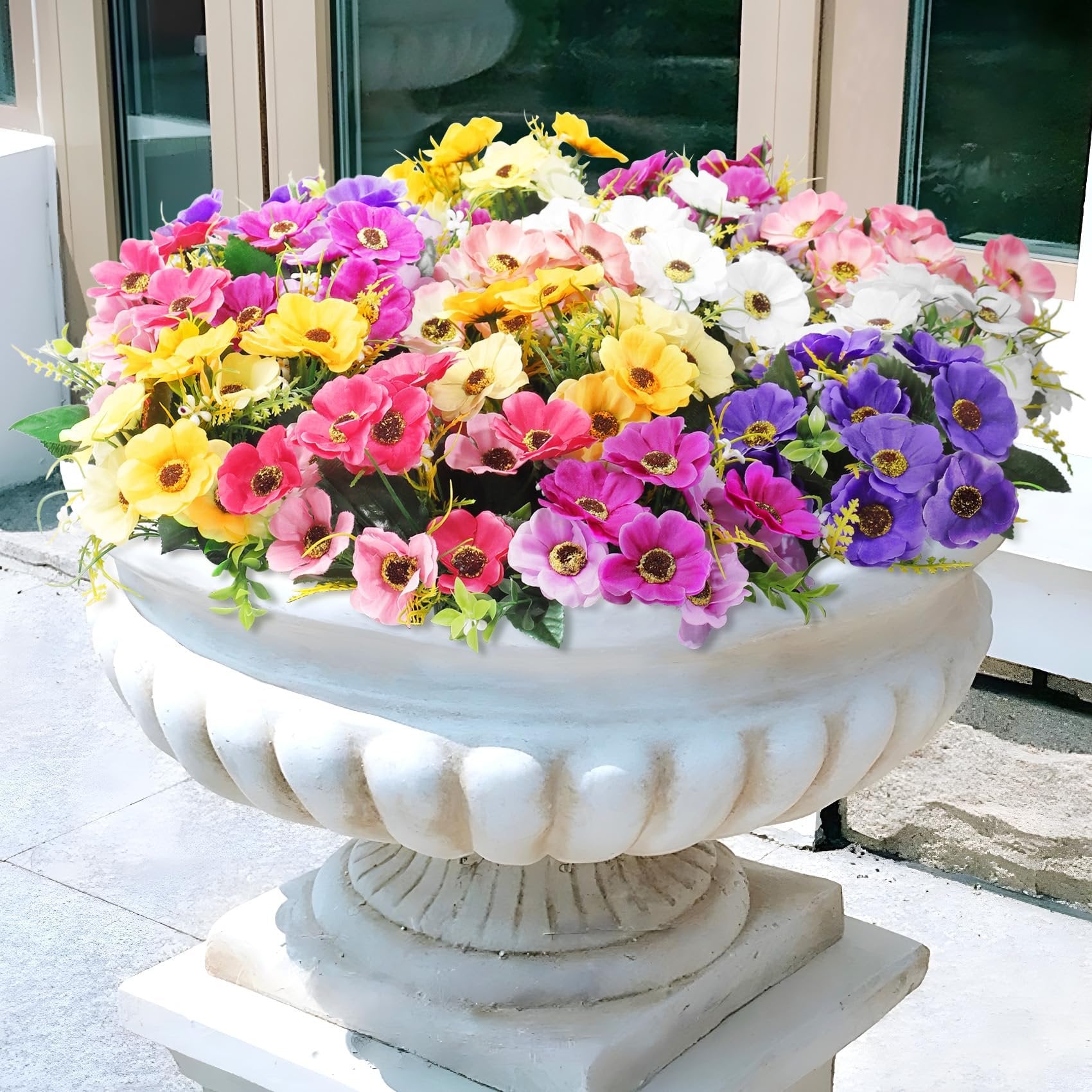 A large white urn filled with a vibrant mix of artificial daisy silk flowers in pink, yellow, purple, and white.